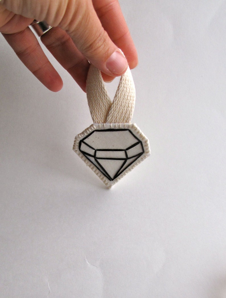 May include: A diamond-shaped, white and black graphic design with a cream-colored ribbon loop. The diamond design has a black outline and internal lines, creating a geometric pattern. The ribbon is held up by a hand.