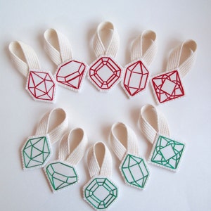 May include: A collection of ten fabric gemstone ornaments with cream-colored ribbon loops. The gemstones are white with red or green embroidered outlines. The shapes include diamonds, emeralds, and other geometric designs.