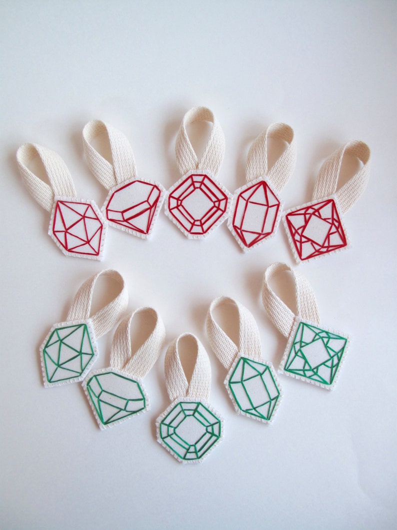 May include: A collection of ten fabric gemstone ornaments with cream-colored ribbon loops. The gemstones are white with red or green embroidered outlines. The shapes include diamonds, emeralds, and other geometric designs.
