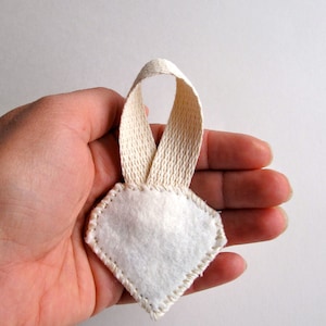 May include: A white felt ornament with a diamond shape and a cream-colored ribbon loop. The edges of the ornament are stitched with cream-colored thread. The ornament is held in a person's hand.