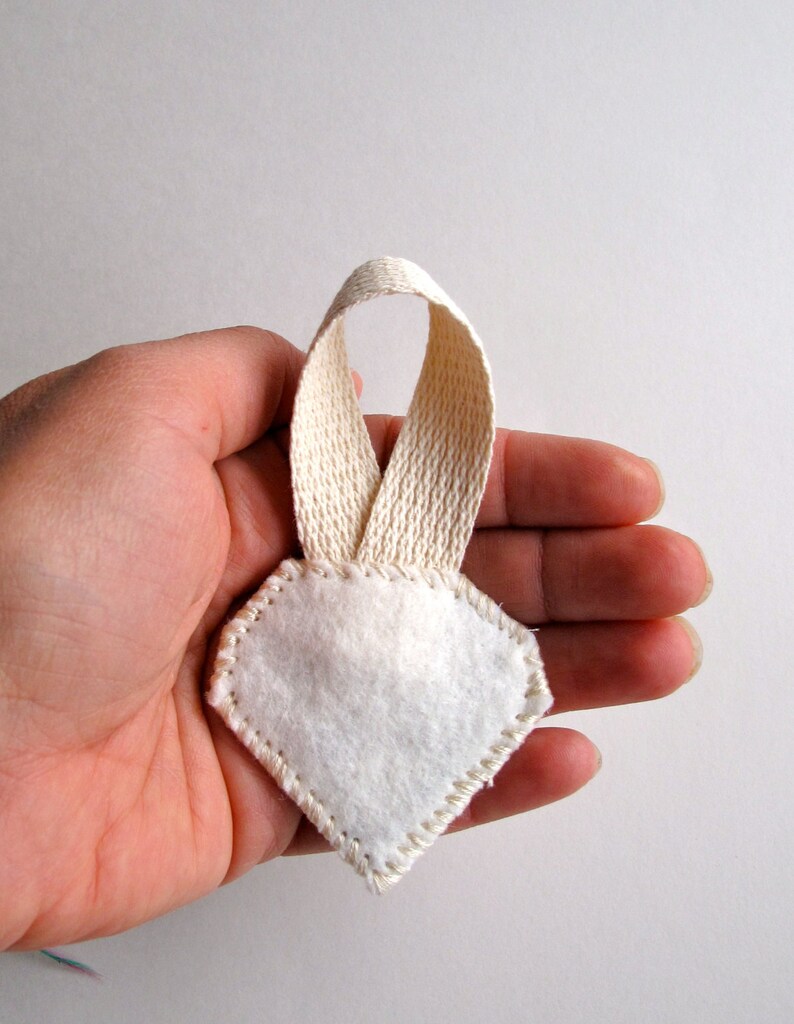 May include: A white felt ornament with a diamond shape and a cream-colored ribbon loop. The edges of the ornament are stitched with cream-colored thread. The ornament is held in a person's hand.