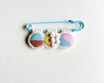 Hand embroidered pin for sweaters with large blue safety pin pink blue tan yellow threads and shell beads on cream muslin An Astrid Endeavor