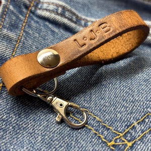 May include: A tan leather keychain loop featuring a silver-toned metal snap button and a silver lobster clasp. The leather strap is embossed with the letters L, J, and B, separated by small dots. The item is constructed from a single strip of leather.