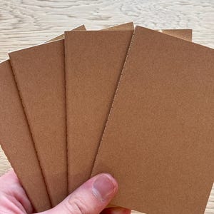 May include: A set of four small, rectangular notebooks with plain, light brown kraft paper covers. Each notebook features a simple stitched spine along the left edge, holding the pages together in a clean, minimalist, and functional design.