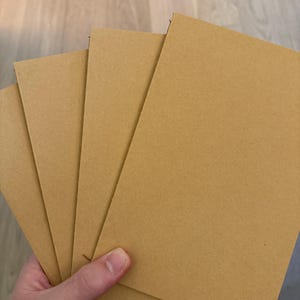 May include: A set of four rectangular cards made of light brown, smooth, matte-finish paper stock. The cards are stacked in a fan-like arrangement, displaying a uniform tan color and clean, sharp edges on all sides of the paper material.