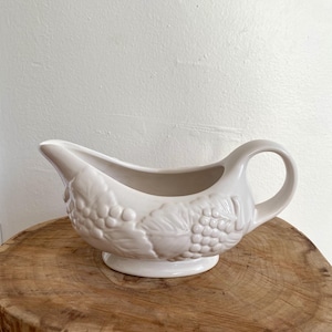 May include: Cream-coloured ceramic gravy boat with a handle and spout. The boat has a raised grape and leaf design on the side. It stands on a small, round base. The gravy boat is on a wooden surface.