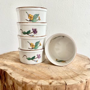 Puede incluir: Cinco ramequines de cerámica blanca apilados, cada uno con un diseño floral de flores amarillas y moradas, hojas verdes y bordes marrones. Un ramequín está separado, mostrando el interior.