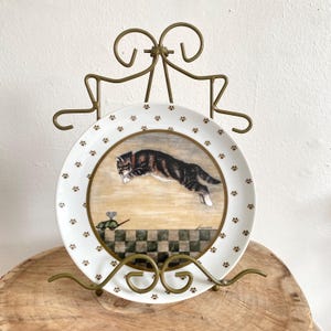 May include: A decorative plate featuring a cat leaping towards a toy mouse. The plate has a white background with brown paw prints around the edge. The plate is displayed on a gold-colored metal stand.