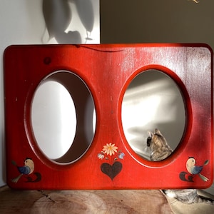May include: A rectangular, red wooden frame with two oval openings, possibly for mirrors. The frame is decorated with painted birds, flowers, and a heart. The frame is resting on a wooden surface.