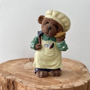 Puede incluir: Una figura de oso pardo vestida de chef, con gorro blanco, camisa verde y delantal amarillo claro. El oso sostiene un pastel pequeño en una pata y una cuchilla en la otra. La figura está sobre una superficie de madera.