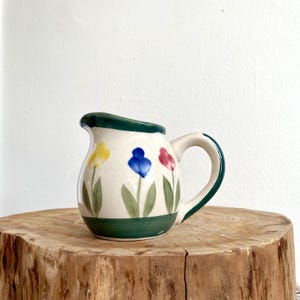 Könnte beinhalten: Ein kleiner, cremefarbener Keramikkrug mit grünem Rand und Griff. Der Krug ist mit bunten Tulpenmotiven in Gelb, Blau und Rot verziert. Der Boden des Kruges hat ein grünes Band.
