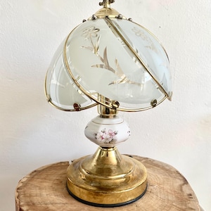 Peut inclure: Une lampe de table vintage en laiton avec un abat-jour en verre dépoli orné de motifs floraux et d'oiseaux gravés. La lampe a une base en céramique blanche ornée de motifs floraux roses. La base de la lampe est ronde et en laiton.