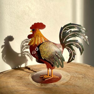 May include: A metal rooster sculpture with a red comb, yellow beak, and green tail feathers. The rooster has a rusty red body with yellow and green accents. It stands on a circular base with yellow legs. The rooster's shadow is visible on the wall.