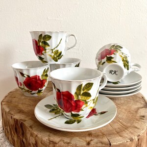 Puede incluir: Juego de tazas y platillos de té de porcelana blanca con un diseño de rosa roja y hojas verdes. Las tazas tienen un borde festoneado y ribete dorado. Los platillos están apilados.