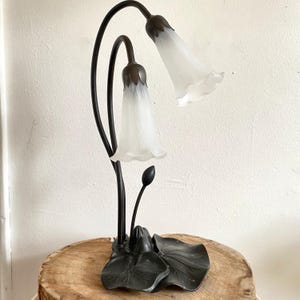 May include: A decorative table lamp with a floral design. The lamp features two white, bell-shaped shades and a black, curved metal frame. The base is a stylized leaf design. The lamp is placed on a wooden surface.