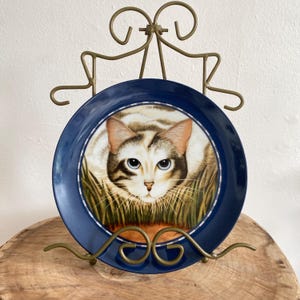 May include: A decorative plate featuring a cat with blue eyes, set against a backdrop of grass. The plate has a dark blue border with a white inner ring. The plate is displayed on a gold-colored metal stand.