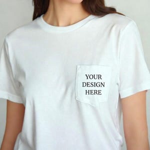 May include: White t-shirt with a pocket on the chest. The pocket features the text "YOUR DESIGN HERE" in black. The shirt has short sleeves and a crew neck.
