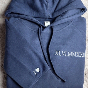 May include: Navy blue hoodie with a drawstring hood. The left sleeve cuff features a white heart and the letter "J". The front of the hoodie displays the Roman numerals "XI.VI.MMXXII" in white. The hoodie is made of a soft material.