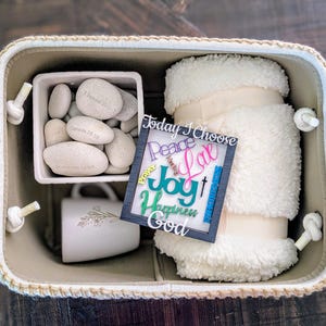 May include: A beige woven basket filled with a white fluffy blanket, a small white square container of stones, a white mug with a floral design, and a framed sign that reads "Today I Choose Peace, Love, Joy, Happiness, God."
