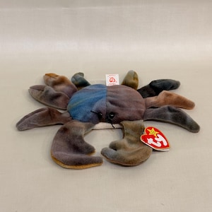 Könnte beinhalten: Ein Plüschtier in Krabbenform mit einem Batikmuster in Blau, Braun und Grau. Das Stofftier hat acht Beine, zwei große Scheren und kleine schwarze Augen. Ein rotes, herzförmiges Ty-Etikett ist an einer der Scheren befestigt, ein weißes Etikett hinten.
