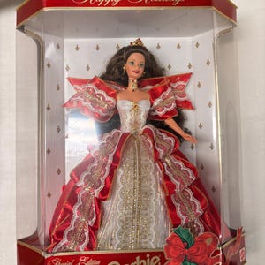 Puede incluir: Una muñeca Barbie en una vitrina transparente. La muñeca lleva un vestido rojo y blanco con detalles dorados y una corona. La vitrina tiene un borde rojo con "Happy Holidays" y "Special Edition Barbie".