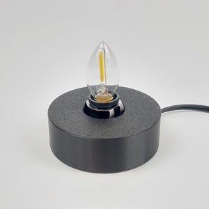 May include: A black, cylindrical lamp base with a textured surface, holding a clear, teardrop-shaped light bulb. The bulb has a yellow filament. A black power cord extends from the base.