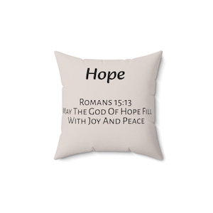 Op de afbeelding: Een vierkant sierkussen in een lichtbeige kleur met het woord Hope in zwarte tekst, gevolgd door de bijbelverwijzing Romans 15:13 en de tekst May The God Of Hope Fill With Joy And Peace gedrukt in een eenvoudig zwart schreefloos lettertype.