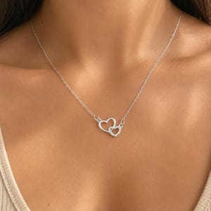 May include: A silver-toned necklace featuring two interlocking heart-shaped pendants. One heart is a plain polished metal outline, while the smaller heart is encrusted with small clear stones. The pendant hangs from a delicate, thin silver-toned chain.