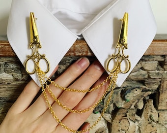 May include: Two gold-toned scissor-shaped brooches with a chain attached to each. The brooches are pinned to a white collared shirt.