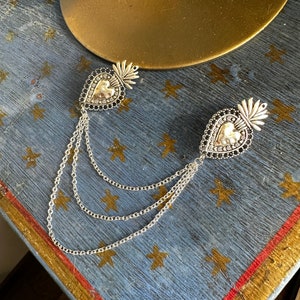 May include: Two silver heart-shaped charms with black accents and a beaded border, connected by three silver chains.