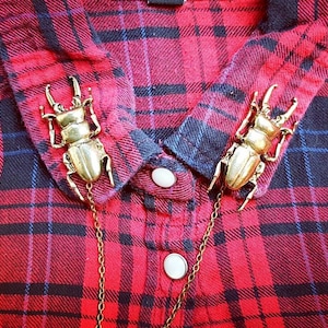 May include: A red and black plaid collared shirt featuring two gold-toned beetle-shaped collar pins connected by a thin gold-toned metal chain. The shirt has white buttons down the front placket and a pointed collar design with visible fabric texture.