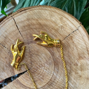 May include: Two gold-colored hand-shaped brooches connected by a delicate chain. The brooches are detailed with intricate finger and wrist designs. The jewelry rests on a natural wood surface, with green foliage in the background.