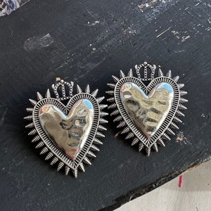 Könnte beinhalten: Zwei silberne herzförmige Anhänger mit einem Kronendesign und einem stacheligen Rand. Die Anhänger befinden sich auf einer schwarzen Oberfläche.