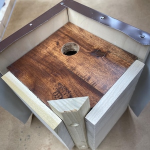 May include: A wooden birdhouse with a brown stained interior and a natural wood exterior. The birdhouse has a square shape with a circular entrance hole and a metal roof trim. The birdhouse is designed for outdoor use.