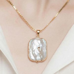 Puede incluir: Un collar dorado con un colgante rectangular con una gran perla blanquecina. La perla está engastada en un marco dorado y cuelga de una fina cadena dorada. El collar se muestra sobre un fondo blanco.