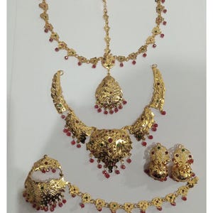 May include: A gold-coloured jewellery set comprising a necklace, earrings, headpiece, and a decorative piece. The set is embellished with red and green accents. The jewellery has an ornate design, displayed on a white surface.