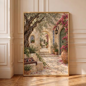 Op de afbeelding: Een ingelijste kunstdruk met een mediterrane binnenplaats met een grote olijfboom, stenen bogen, een groene houten deur en felroze bougainvillea-bloemen. De scène bevat terracotta potten, een kussen met patroon en een lantaarn.