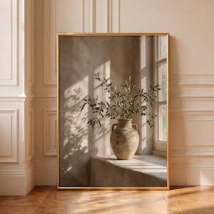 Könnte beinhalten: Ein rechteckiges Wandbild in einem dünnen goldenen Rahmen zeigt eine Fotografie einer strukturierten beigen Keramikvase mit zwei Henkeln. Die Vase enthält einen grünen Olivenzweig und steht auf einem Sims an einem Fenster mit Schattenwurf.