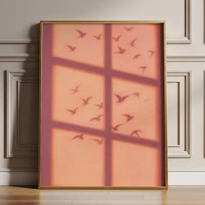 Op de afbeelding: Een ingelijste kunstdruk met een perzikkleurige achtergrond en silhouetten van vogels in vlucht. De schaduw van een raam creëert een rasterpatroon. De lijst is goudkleurig.