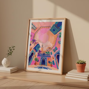 May include: A framed art print depicting a vibrant, stylized courtyard scene. The artwork features a pink and orange sky, blue archways, and colorful details. The print is displayed in a light-colored wooden frame, leaning against a neutral wall.