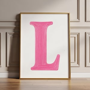 Puede incluir: Impresión enmarcada con una gran letra "L" de color rosa vibrante. La letra está pintada con pinceladas visibles, sobre un fondo blanco. El marco dorado añade un toque de elegancia, ideal para la decoración del hogar.