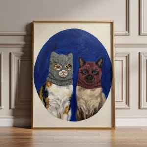 以下が含まれることがあります： 青い背景に、ニットのバラクラバをかぶった2匹の猫が描かれた額入りの絵画。1匹は三毛猫、もう1匹はシャム猫です。アートワークは金のフレームに入っており、木の床に置かれています。