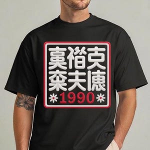 Pode incluir: Camiseta preta com um design gráfico quadrado branco e vermelho contendo caracteres asiáticos e o número "1990". O design gráfico é delineado em vermelho e branco. A camiseta é de manga curta.