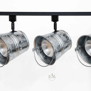 Może przedstawiać: Trzy srebrne metalowe lampy wiszące w kształcie wiadra wiszą na czarnym torze. Każda lampa ma przezroczystą żarówkę i srebrny metalowy uchwyt.