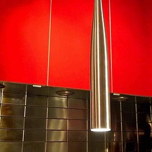 May include: A silver baseball bat-shaped pendant light fixture hangs from a ceiling against a red background. The light is on and illuminates the surrounding area.