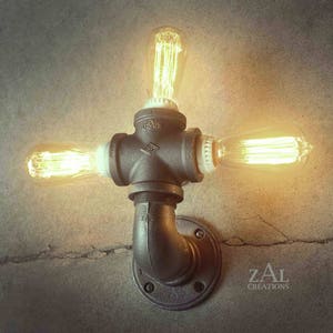 May include: A wall-mounted light fixture made from black industrial pipe with three Edison light bulbs. The fixture is mounted on a gray wall with the text "ZAL CREATIONS" in the bottom right corner.
