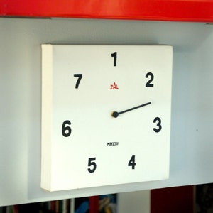 May include: A white clock face with black numbers 1 through 7 and black hands. The clock face also has the text "ZAL" in red and "MMXIII" in black. The clock is mounted on a white wall.