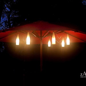 May include: A red patio umbrella with a string of lights made from wine bottles hanging from it. The lights are turned on and illuminating the area below. The umbrella is in a dark setting with trees in the background.