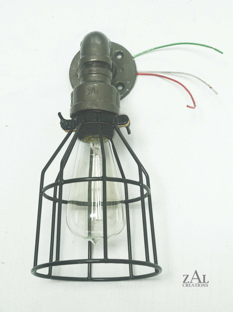 May include: A black metal cage light fixture with a clear glass bulb. The fixture is attached to a gray metal pipe and has a round base with wires coming out of it. The text "ZAL CREATIONS" is visible in the bottom right corner of the image.