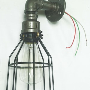 May include: A black metal cage light fixture with a clear glass bulb. The fixture is attached to a gray metal pipe and a round wall mount. The wires are visible and are red, green, and white.